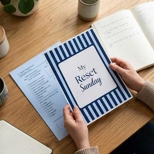 May include: A paper planner cover with a dark blue and white vertical striped border and a white centre rectangle featuring the text My Reset Sunday in dark blue font. A light blue checklist page with printed text is visible underneath the cover.