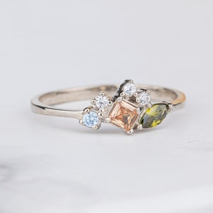 May include: A delicate silver ring featuring a cluster of small clear and light blue gemstones, a larger square peach-coloured gemstone, and two green marquise-shaped gemstones.