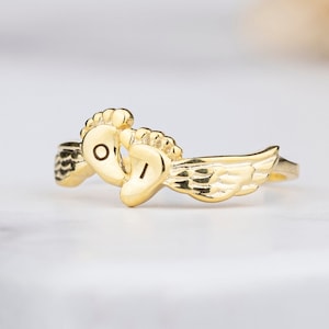 以下が含まれることがあります： 天使の羽根と赤ちゃんの足跡がついた金の指輪。足跡には「O」と「I」の文字が刻まれています。