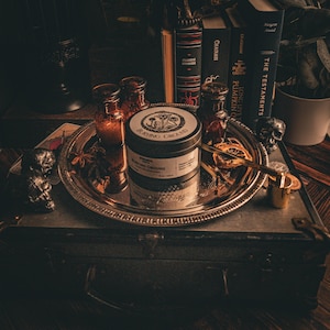 May include: A black metal tin with the words "Burning Ground" printed on the front. The tin is sitting on a silver tray with a variety of items, including a skull, a book, and a bottle of liquid. The tin is surrounded by a variety of other items, including a book, a bottle of liquid, and a skull.