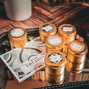May include: Six gold candle tins with various designs and labels. The labels include "Fall of the Patriarchy", "Memento Mori", "Golden Age", and "Burlesque". The candles are arranged on a black and gold tray with a card that reads "FAVORITES CANDLE DISCOVERY COCONUT WAX BLEND SCENT"