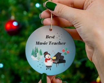 May include: A blue and white Christmas ornament with a snowman playing a piano. The ornament says "Best Music Teacher" on it.