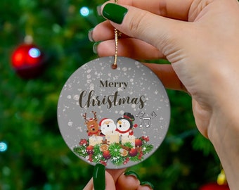 May include: A round Christmas ornament with a gray background and the words "Merry Christmas" in script. The ornament features cartoon characters of a reindeer, Santa, and a snowman. The ornament is held by a person.