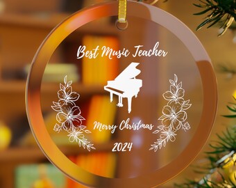 May include: A clear glass Christmas ornament with a white inscription that reads "Best Music Teacher Merry Christmas 2024". The ornament features a white silhouette of a grand piano with floral accents.