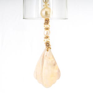 May include: A delicate, pearlescent shell pendant hangs from a gold chain with small white and gold beads. The pendant is shaped like a teardrop and has a subtle iridescent sheen.