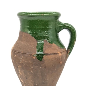 May include: A green and brown ceramic jug with a handle. The jug has a worn, rustic look with some of the green glaze chipped off.