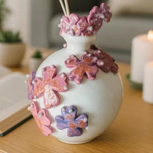 May include: A white ceramic vase decorated with pink, purple, and orange floral designs and faux pearls. The vase contains two blue flower decorations on wooden sticks. The vase is on a wooden table.