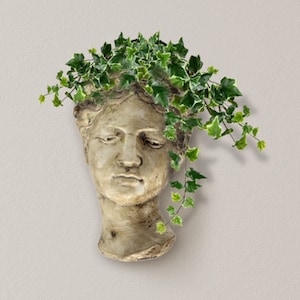 Op de afbeelding: Een witte gipsen buste plantenpot met een gebeeldhouwd gezicht en een kroon van groene klimop planten.