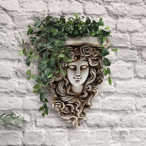 Op de afbeelding: Een witte gipsen wandsculptuur van een vrouwenhoofd met golvend haar, omringd door groene, bladerige planten in een hangende pot. De sculptuur is bevestigd aan een witte bakstenen muur.