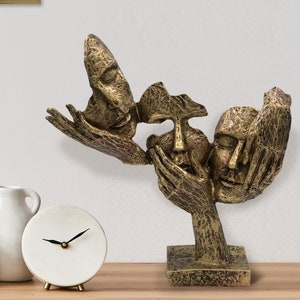 May include: A bronze-effect sculpture depicting three faces, with hands partially covering them. The sculpture has a textured finish and is mounted on a square base. A decorative art piece.