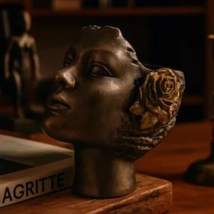 Puede incluir: Una escultura de color bronce de la cara de una mujer con un detalle de rosa en el lateral. La escultura está colocada sobre una superficie de madera, con libros y otros objetos decorativos en el fondo. Es un objeto decorativo.