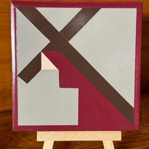 May include: A small, square abstract painting in shades of burgundy, brown, gray, and beige, mounted on a miniature wooden easel. The artwork features geometric shapes and lines, creating a modern, minimalist design.
