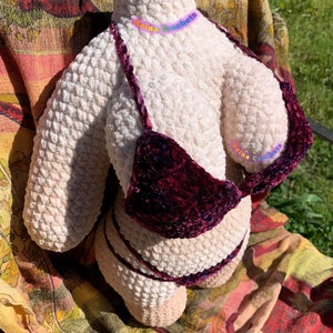 May include: A crocheted plush doll wearing a burgundy velvet bikini. The doll is sitting on a patterned fabric.