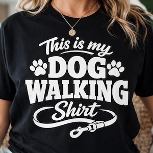 Könnte beinhalten: Schwarzes T-Shirt mit dem weißen Text "This is my DOG WALKING Shirt". Das Design beinhaltet Pfotenabdrücke und eine Leinen-Grafik. Eine goldene Halskette ist sichtbar.