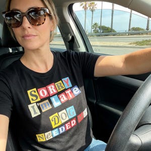Puede incluir: Camiseta negra de cuello redondo con el texto "SORRY I'm Late DOG NEEDED ME" en letras coloridas estilo recortado. La persona de la imagen lleva gafas de sol y está en un coche.