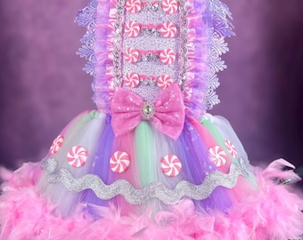 May include: A colorful, candy-themed dress with a bodice adorned with faux candies and a large pink bow. The skirt features layers of rainbow-colored tulle and is trimmed with a fluffy pink feather boa. Silver accents and more candy details complete the whimsical design.