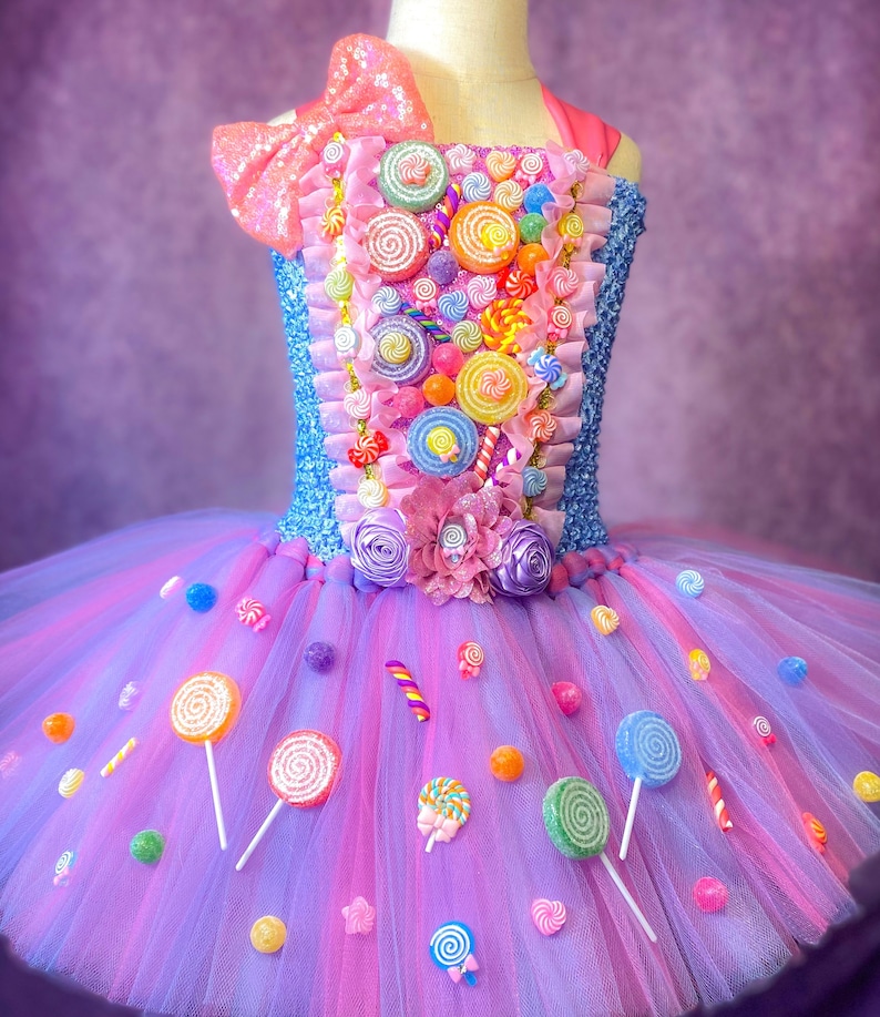 May include: A purple and pink tulle tutu with a candy theme. The tutu is decorated with colorful lollipops, candy canes, and gumdrops. The bodice of the tutu is blue and pink and is covered in colorful candy decorations.