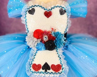 May include: A blue tulle tutu with a white bodice featuring a playing card design and red and black hearts and spades. The bodice is decorated with a flower arrangement of red, white, blue, and black flowers.