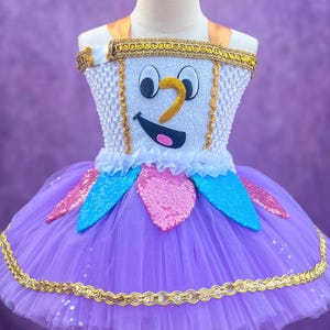 May include: A whimsical tutu dress featuring a cartoon teacup design. The bodice is white with a smiling face, gold trim, and a yellow handle. The skirt is layered purple tulle, accented with sequined details.