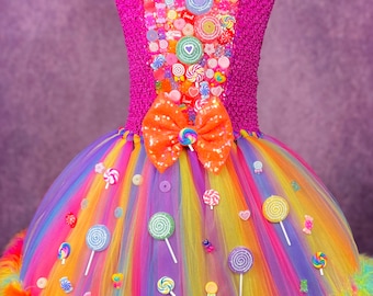 Könnte beinhalten: Ein farbenfrohes Tutu-Kleid mit Candy-Thema. Das Oberteil in Magenta ist strukturiert und mit verschiedenen Süßigkeiten verziert. Eine große orangefarbene Pailletten-Schleife sitzt an der Taille. Der Regenbogen-Tüllrock ist mit Lollis und Candy-Akzenten verziert, mit bunten Federn besetzt.