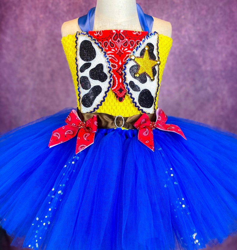 Puede incluir: Un tut&uacute; de tul azul con un corpi&ntilde;o de tem&aacute;tica vaquera amarillo y rojo. El corpi&ntilde;o presenta un pa&ntilde;uelo rojo, una estrella de sheriff y un dise&ntilde;o de estampado de vaca. El tut&uacute; est&aacute; decorado con lentejuelas plateadas.