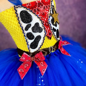 Puede incluir: Un tut&uacute; de tul azul y amarillo con un lazo de bandana rojo y blanco. La parte superior del tut&uacute; es un top de crochet amarillo con estampado de vaca y dise&ntilde;o de bandana rojo.