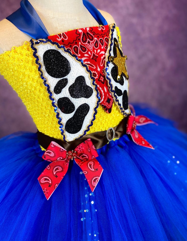 Puede incluir: Un tut&uacute; de tul azul y amarillo con un lazo de bandana rojo y blanco. La parte superior del tut&uacute; es un top de crochet amarillo con estampado de vaca y dise&ntilde;o de bandana rojo.
