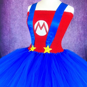 Pode incluir: Um vestido de tutu vermelho e azul com um "M" branco e estrelas amarelas. O vestido &eacute; feito de tule e tem um corpete de croch&ecirc;.