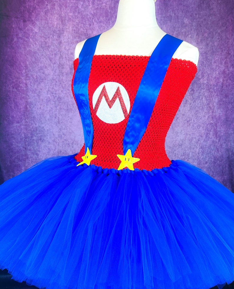 Pode incluir: Um vestido de tutu vermelho e azul com um "M" branco e estrelas amarelas. O vestido &eacute; feito de tule e tem um corpete de croch&ecirc;.