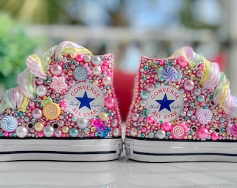 Könnte beinhalten: Ein Paar weiße Converse-Sneaker mit hohem Schaft, verziert mit bunten Süßigkeiten, Perlen und Strasssteinen. Die Sneaker haben eine rosa Basis und ein Regenbogen-Swirl-Design an den Schnürsenkeln.