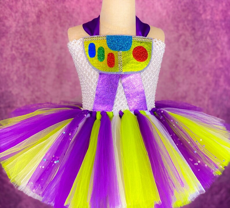 May include: A white tulle tutu with purple, yellow, and white stripes. The tutu has a purple and white bodice with a colorful toy character design.
