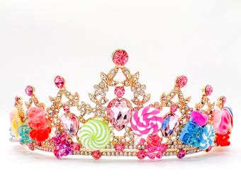 Könnte beinhalten: Eine goldene Krone mit rosa, weißen und grünen Süßigkeiten-Dekorationen. Die Krone ist mit Strasssteinen verziert und zeigt verschiedene Süßigkeitenformen, darunter Lutscher, Gummibärchen und Zuckerstangen.
