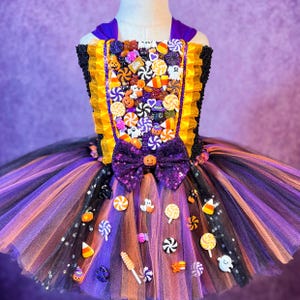 May include: A Halloween-themed tutu dress with a bodice adorned with candy-shaped embellishments. The dress features a purple bow with a pumpkin accent, a yellow and black bodice, and a layered skirt in black, purple, and orange tulle.
