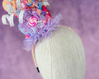 Könnte beinhalten: Ein verspieltes Stirnband mit einem bunten Süßigkeiten-Thema. Es verfügt über eine Vielzahl von bunten Lutschern, Bändern und Blumen. Das Stirnband besteht aus einem weißen Gummiband und ist perfekt, um jedem Outfit einen Hauch von Spaß zu verleihen.