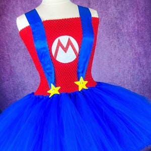 Pode incluir: Um vestido de tutu azul e vermelho com um corpete vermelho que apresenta um "M" branco e al&ccedil;as azuis. A saia &eacute; feita de tule e tem detalhes de estrelas amarelas.