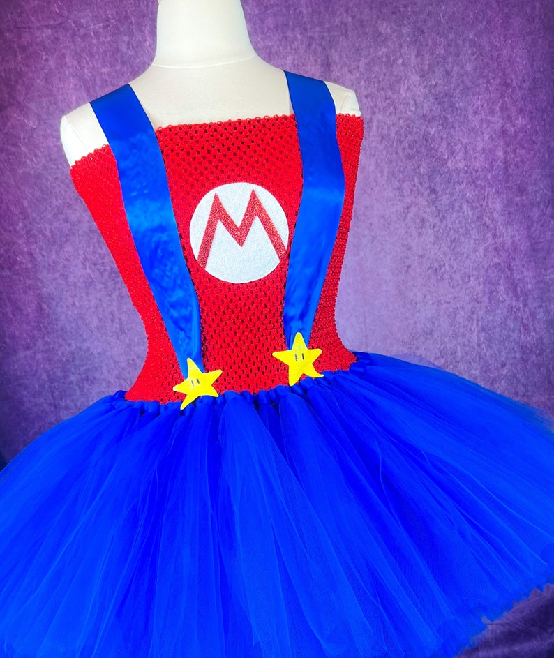 Pode incluir: Um vestido de tutu azul e vermelho com um corpete vermelho que apresenta um "M" branco e al&ccedil;as azuis. A saia &eacute; feita de tule e tem detalhes de estrelas amarelas.