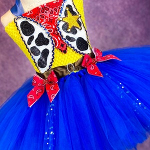 Puede incluir: Un tut&uacute; de tul azul con un corpi&ntilde;o amarillo y rojo con una estrella de vaquero, un pa&ntilde;uelo y una impresi&oacute;n de vaca. El corpi&ntilde;o tiene un cintur&oacute;n marr&oacute;n con una hebilla plateada. El tut&uacute; est&aacute; decorado con purpurina plateada.