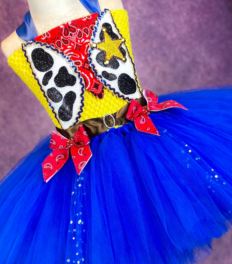 Puede incluir: Un tut&uacute; de tul azul con un corpi&ntilde;o amarillo y rojo con una estrella de vaquero, un pa&ntilde;uelo y una impresi&oacute;n de vaca. El corpi&ntilde;o tiene un cintur&oacute;n marr&oacute;n con una hebilla plateada. El tut&uacute; est&aacute; decorado con purpurina plateada.