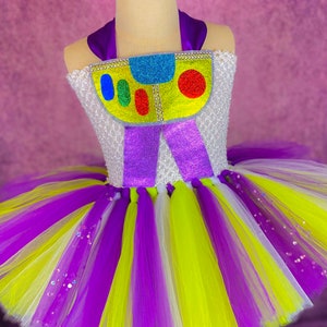 May include: A purple, white, and yellow tulle tutu with a white bodice featuring a colorful cartoon character with the text "Buzz Lightyear".