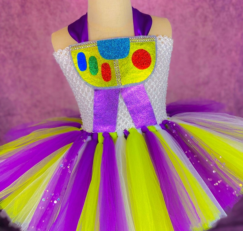 May include: A purple, white, and yellow tulle tutu with a white bodice featuring a colorful cartoon character with the text "Buzz Lightyear".