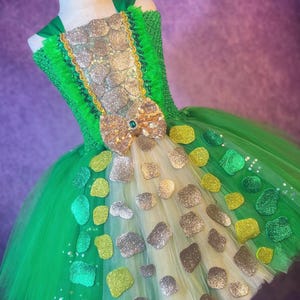May include: A green and gold tutu dress with a bodice adorned with glittery silver and gold embellishments. The dress features a large bow with a green jewel and is decorated with various glittery shapes. The tulle skirt is layered with green and cream fabric.
