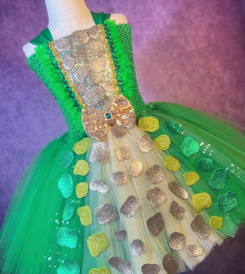 May include: A green and gold tutu dress with a bodice adorned with glittery silver and gold embellishments. The dress features a large bow with a green jewel and is decorated with various glittery shapes. The tulle skirt is layered with green and cream fabric.