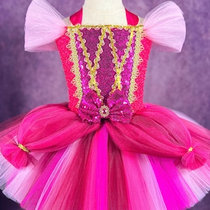 May include: A pink and gold tulle tutu dress with a sequined bodice and a pink bow. The dress has a halter neckline and pink tulle sleeves.
