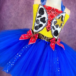 Puede incluir: Un tut&uacute; de tul azul con un corpi&ntilde;o amarillo y rojo con un dise&ntilde;o de vaquero con un pa&ntilde;uelo rojo, una estrella y estampado de vaca. El corpi&ntilde;o tiene un cintur&oacute;n marr&oacute;n con una hebilla.