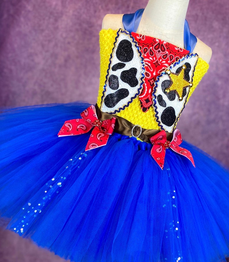Puede incluir: Un tut&uacute; de tul azul con un corpi&ntilde;o amarillo y rojo con un dise&ntilde;o de vaquero con un pa&ntilde;uelo rojo, una estrella y estampado de vaca. El corpi&ntilde;o tiene un cintur&oacute;n marr&oacute;n con una hebilla.