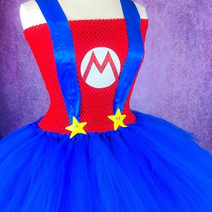 Pode incluir: Um traje de tutu vermelho e azul com um logotipo "M" branco e estrelas amarelas. O traje &eacute; feito de tule e tem um corpete vermelho com al&ccedil;as azuis.