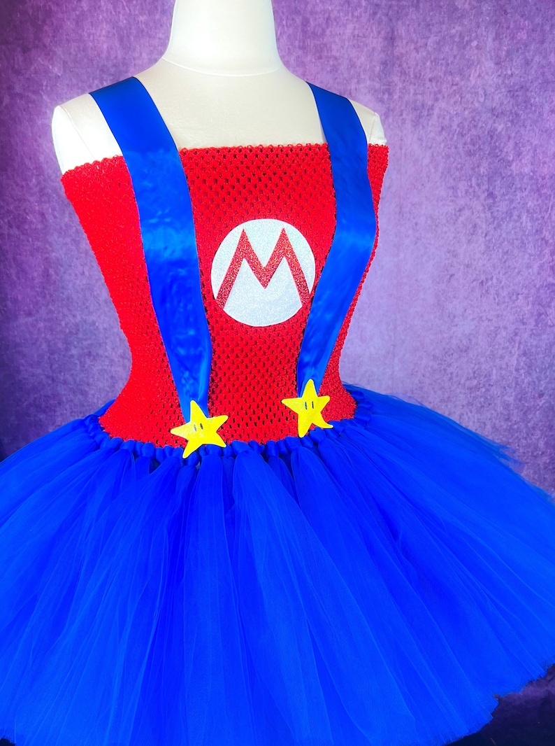 Pode incluir: Um traje de tutu vermelho e azul com um logotipo "M" branco e estrelas amarelas. O traje &eacute; feito de tule e tem um corpete vermelho com al&ccedil;as azuis.