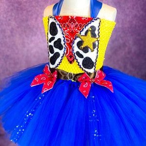 Puede incluir: Un tut&uacute; de tul azul con un chaleco amarillo y rojo con una estrella de sheriff y un patr&oacute;n de bandana. El chaleco tiene un cintur&oacute;n marr&oacute;n con una hebilla plateada.
