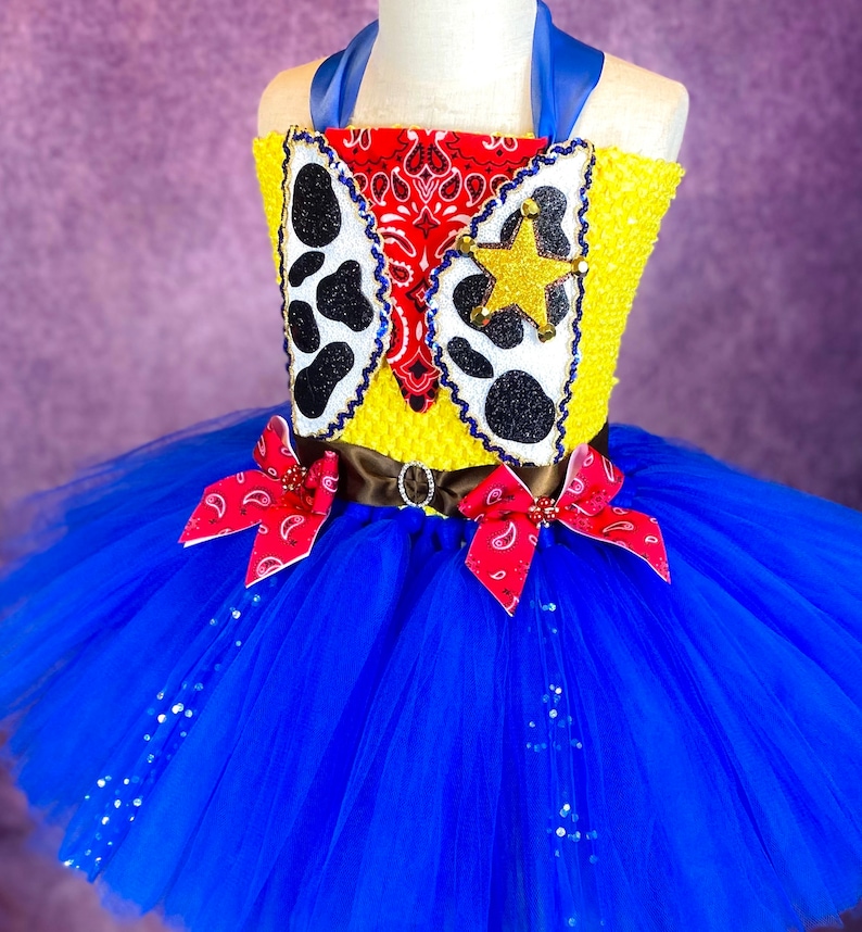 Puede incluir: Un tut&uacute; de tul azul con un chaleco amarillo y rojo con una estrella de sheriff y un patr&oacute;n de bandana. El chaleco tiene un cintur&oacute;n marr&oacute;n con una hebilla plateada.