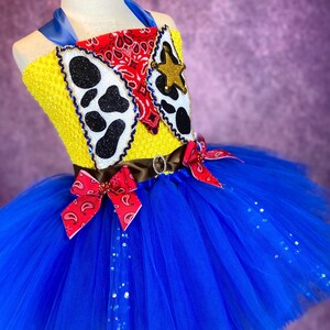 Puede incluir: Un tut&uacute; de tul azul con un corpi&ntilde;o amarillo y rojo con estampado de vaca, un pa&ntilde;uelo rojo y una estrella de sheriff. El corpi&ntilde;o est&aacute; decorado con purpurina.